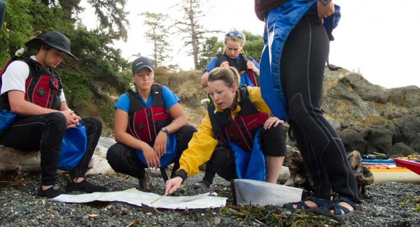 a group of people wearing life jackets examine a map that is spread out on a rocky shore.